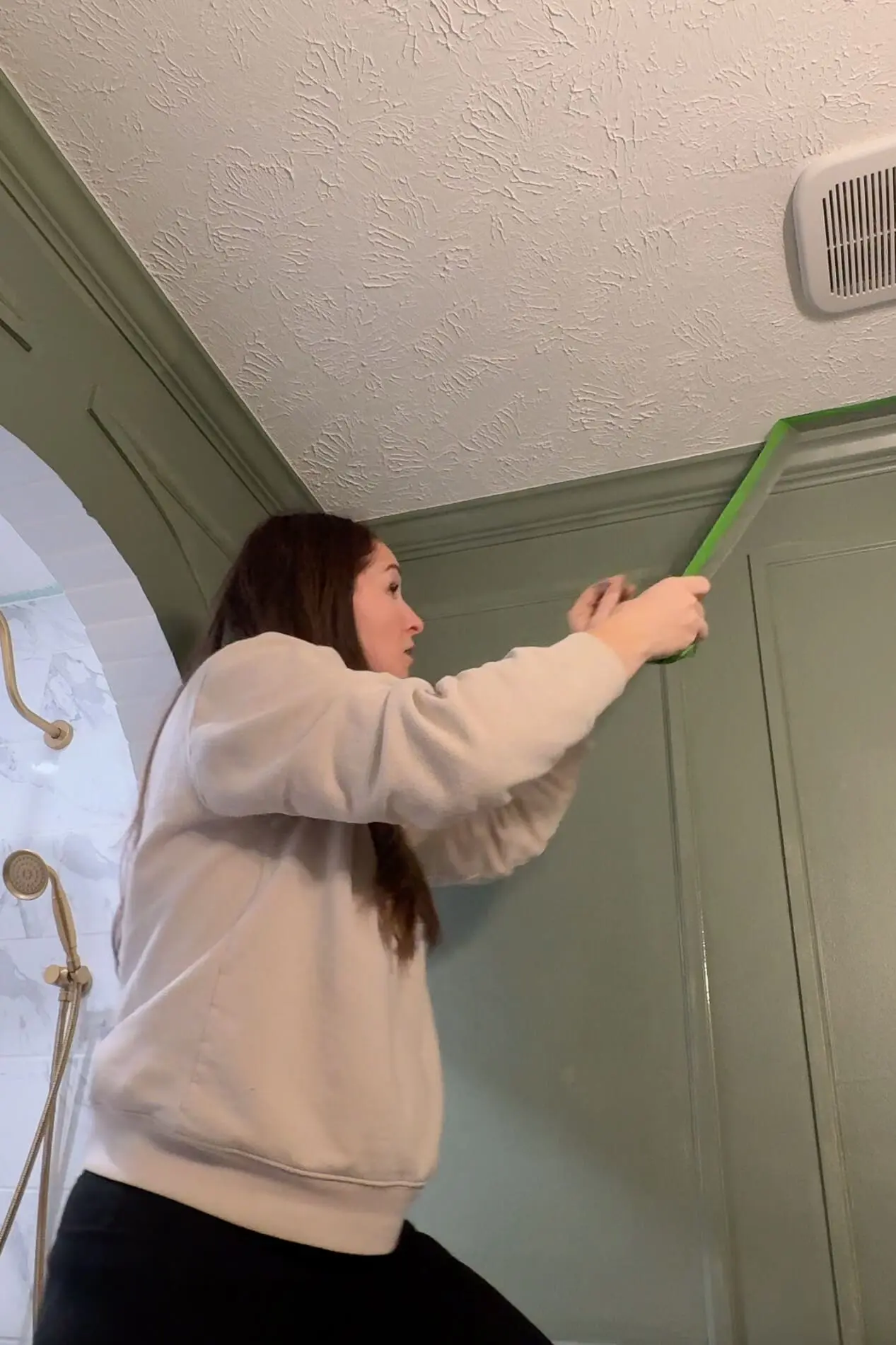 Woman peeling off painter's tape revealing perfectly crisp and clean paint lines at the ceiling edge while painting a dark green bathroom trim accent wall with detailed molding.